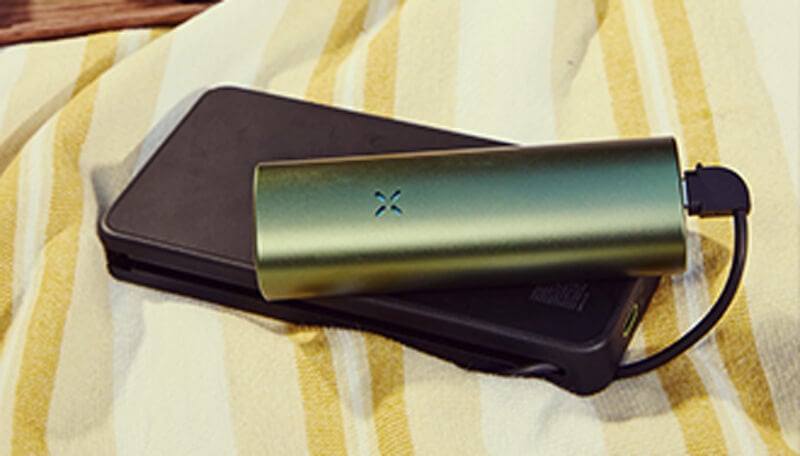 Ricarica del vaporizzatore Pax Flow Un vaporizzatore Pax Flow verde elegante collegato a un caricabatterie portatile nero tramite cavo USB, posizionato su un asciugamano a righe giallo e bianco per una ricarica comoda in movimento