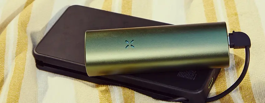 Ricarica del vaporizzatore Pax Flow Un vaporizzatore Pax Flow verde elegante collegato a un caricabatterie portatile nero tramite cavo USB, posizionato su un asciugamano a righe giallo e bianco per una ricarica comoda in movimento.