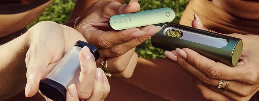 Riempimento del vaporizzatore Pax Flow Due mani che collaborano per riempire il vaporizzatore Pax Flow con cannabis macinato da un piccolo grinder, mostrando il processo di caricamento facile all'aperto su erba verde.