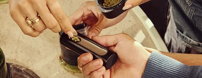 Ricarica del vaporizzatore Pax Flow Una mano che ricarica la camera del vaporizzatore Pax Flow con cannabis macinato fresco usando un piccolo cucchiaino da un grinder, posizionato su un tavolo di marmo per un rifornimento preciso.