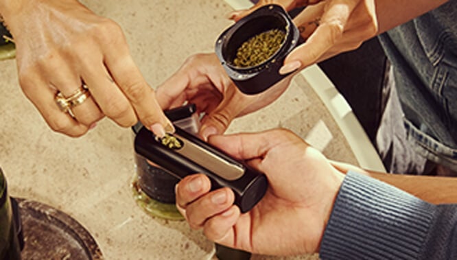 Ricarica del vaporizzatore Pax Flow Una mano che ricarica la camera del vaporizzatore Pax Flow con cannabis macinato fresco usando un piccolo cucchiaino da un grinder, posizionato su un tavolo di marmo per un rifornimento preciso.